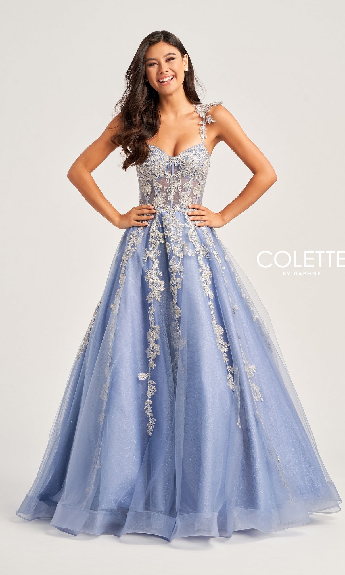 A woman stands smiling against a plain background in the Metallic-Lace Colette Long Tulle Prom Dress CL5165, featuring light blue tulle, floral lace details, and a corset bodice.